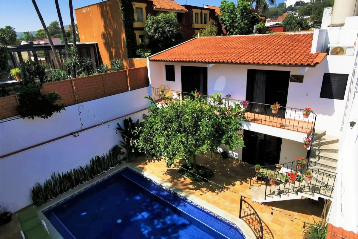 A serene courtyard features a sparkling blue pool surrounded by tiled flooring and lush greenery. Two stories of a building provide a charming backdrop, accented by potted plants. Balconies with wrought iron railings offer views of the tranquil outdoor space.