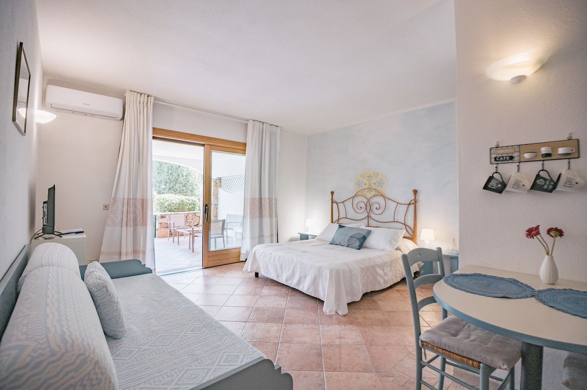 A spacious room features a double bed with bedside tables and lamps. A couch is positioned nearby, offering additional seating. A small dining table with chairs is visible, and natural light enters through the sliding door leading to a furnished patio.