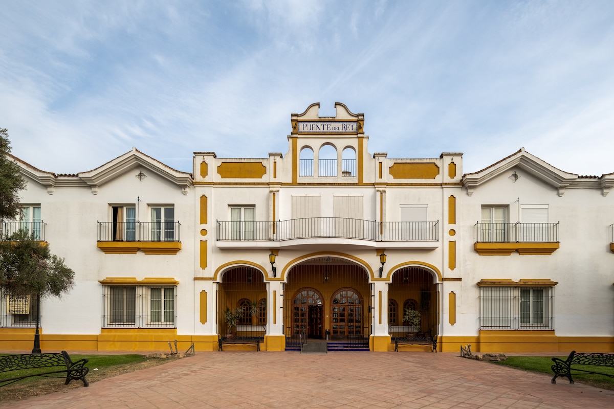 The building features a two-story design with a prominent entrance, characterized by ornate architectural details. A central balcony is flanked by windows, allowing natural light. The exterior is painted in cream and yellow hues, complemented by landscaped grounds and benches in front.