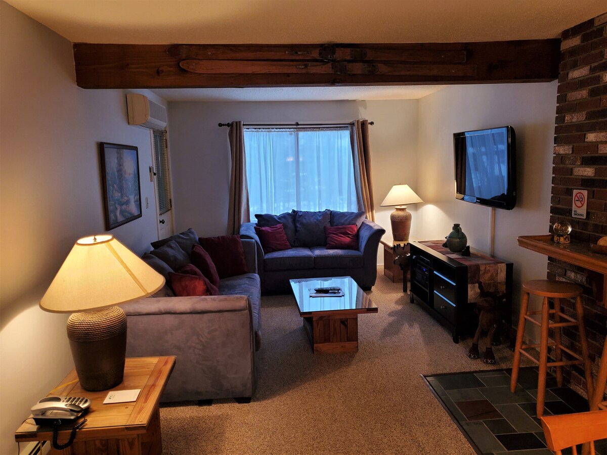 A cozy living room is depicted, featuring a comfortable sectional sofa with cushions in deep red and blue. A glass coffee table is positioned at the center, while a TV is mounted on the wall opposite. Soft light emanates from a lamp beside a well-furnished side table.