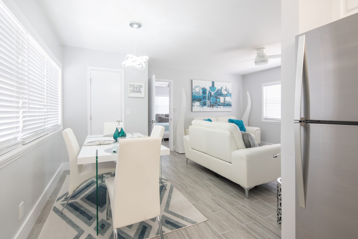 An open-concept living area is showcased with a contemporary white dining table and chairs. A light gray sofa is accented with turquoise cushions, and artwork is displayed on the wall. Natural light streams in through white blinds on the windows.