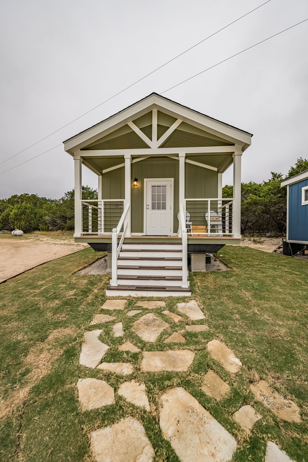 A charming tiny home is featured, showcasing a welcoming front porch with a white railing. The entrance is accentuated by steps leading up to a central front door, framed by a gabled roof. Lush green grass surrounds the stone pathway, contributing to an inviting outdoor space.