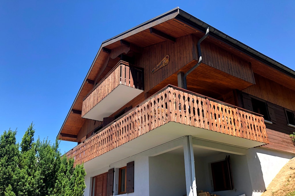 The chalet's exterior showcases a wooden facade with balconies offering views of the surroundings. The structure features multiple levels, complemented by a clear blue sky. Green foliage provides a natural contrast against the building, enhancing the inviting appearance of the property.