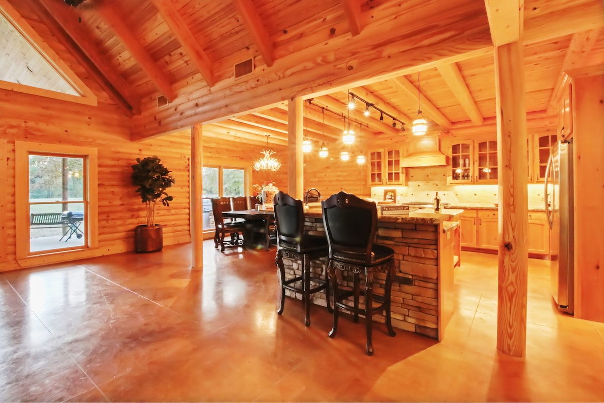 The spacious interior showcases a combination of wooden beams and natural stone accents. An open concept kitchen with stainless steel appliances connects to a dining area, where a rustic wooden table is paired with comfortable black chairs. Large windows provide ample natural light and views of the outdoors.