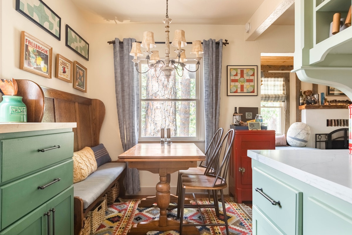 A cozy dining area features a round wooden table surrounded by two black chairs. A cushioned bench lines one side, providing additional seating. Natural light filters through a window adorned with blue curtains, while eclectic artwork decorates the walls, enhancing the inviting ambiance.
