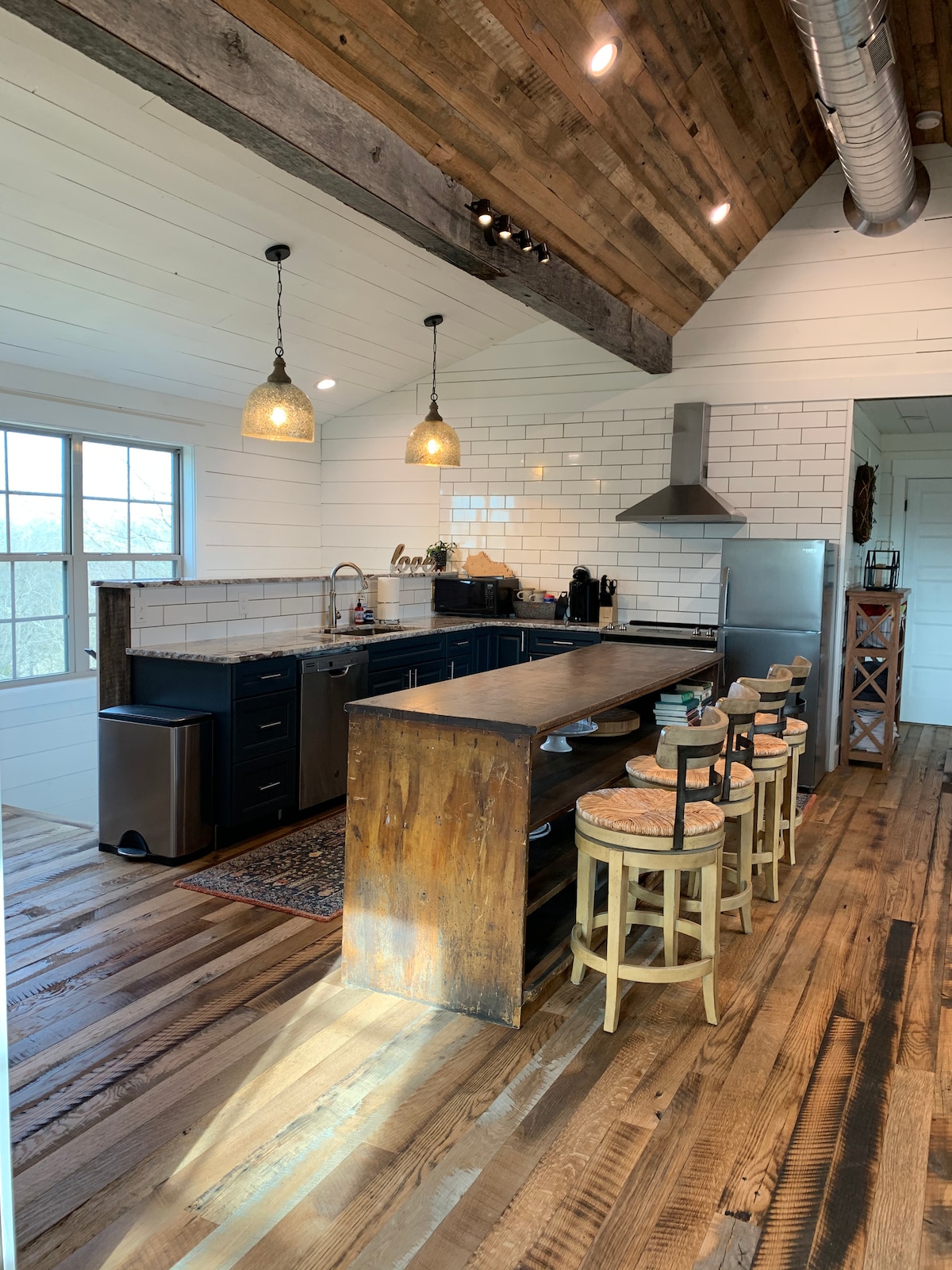 A modern kitchen features dark cabinetry, white subway tile backsplash, and stainless steel appliances. A spacious wooden island provides seating for four with woven stools. Natural light floods in through large windows, highlighting the warm reclaimed wood flooring and vaulted ceiling with rustic beams.