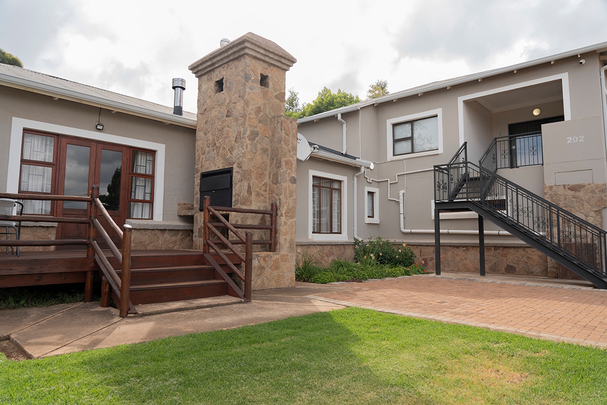 The exterior view of the apartment features a textured stone chimney next to a wooden deck with steps. A paved pathway leads to a parking area, complemented by a manicured lawn and planted greenery. A staircase leads to the upper level of the building.