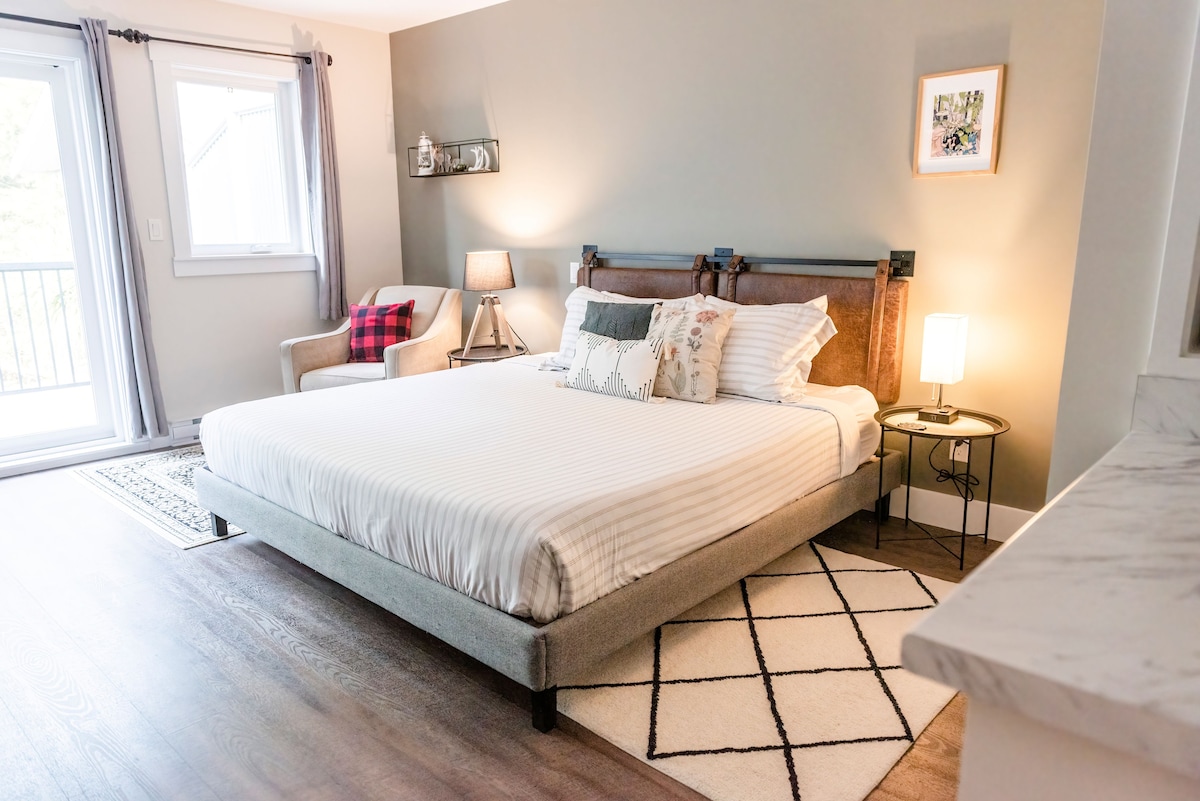 A spacious bedroom features a large, light-colored bed with soft linens and decorative pillows. A cozy accent chair is situated nearby, complemented by a small side table with a lamp. Natural light fills the room through a glass sliding door that opens to a balcony.