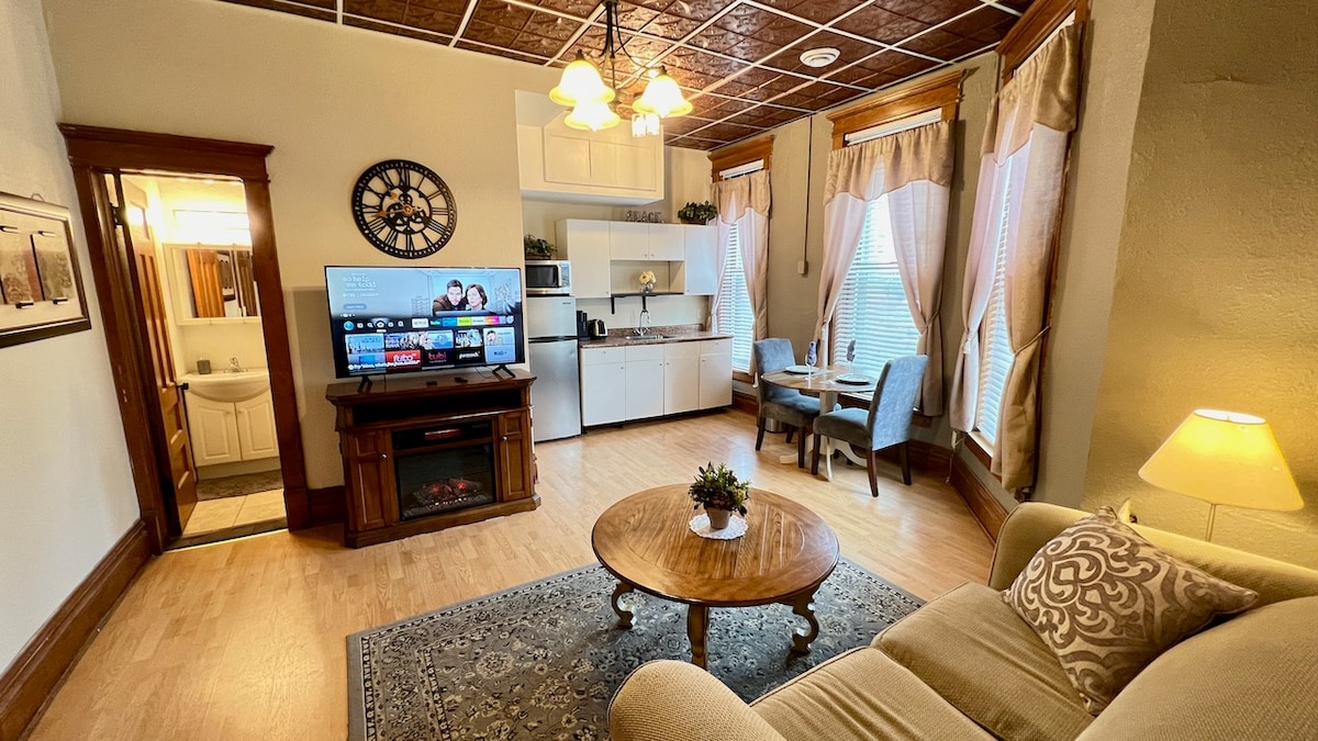 A living area featuring a cozy sofa and a circular wooden coffee table. A flat-screen TV is mounted on a cabinet, displaying various streaming options. Two chairs and a small dining table are situated near the kitchen area, which is equipped with cabinetry and appliances, including a coffee maker.