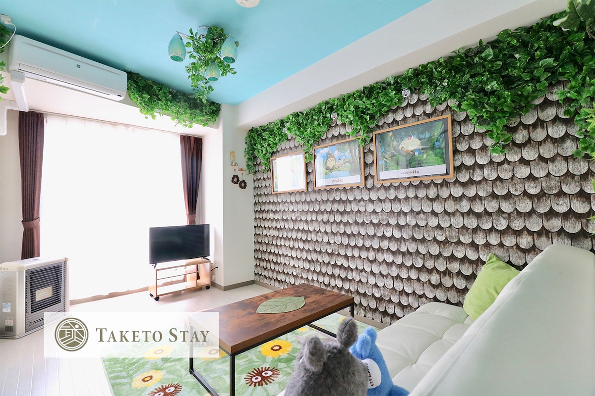 A living area is shown with a soft, white sofa and a decorative rug featuring floral patterns. A wall adorned with leafy greenery adds a natural touch, while framed artworks hang above. A television and air conditioning unit are also visible, enhancing the comfort of the space.