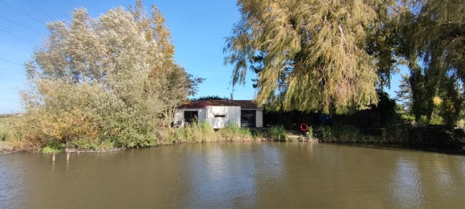 A serene water view highlights a charming accommodation nestled by the water's edge, framed by lush trees and greenery. A glimpse of the building's exterior showcases a simple structure, contributing to the tranquil atmosphere of the surrounding nature.