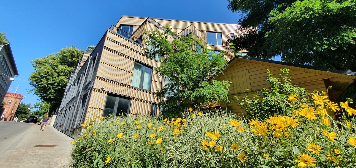 A modern building featuring a mix of materials and large windows is set against a backdrop of vibrant yellow flowers and lush greenery. The exterior is complemented by the neighborhood's architecture, providing a welcoming urban environment.