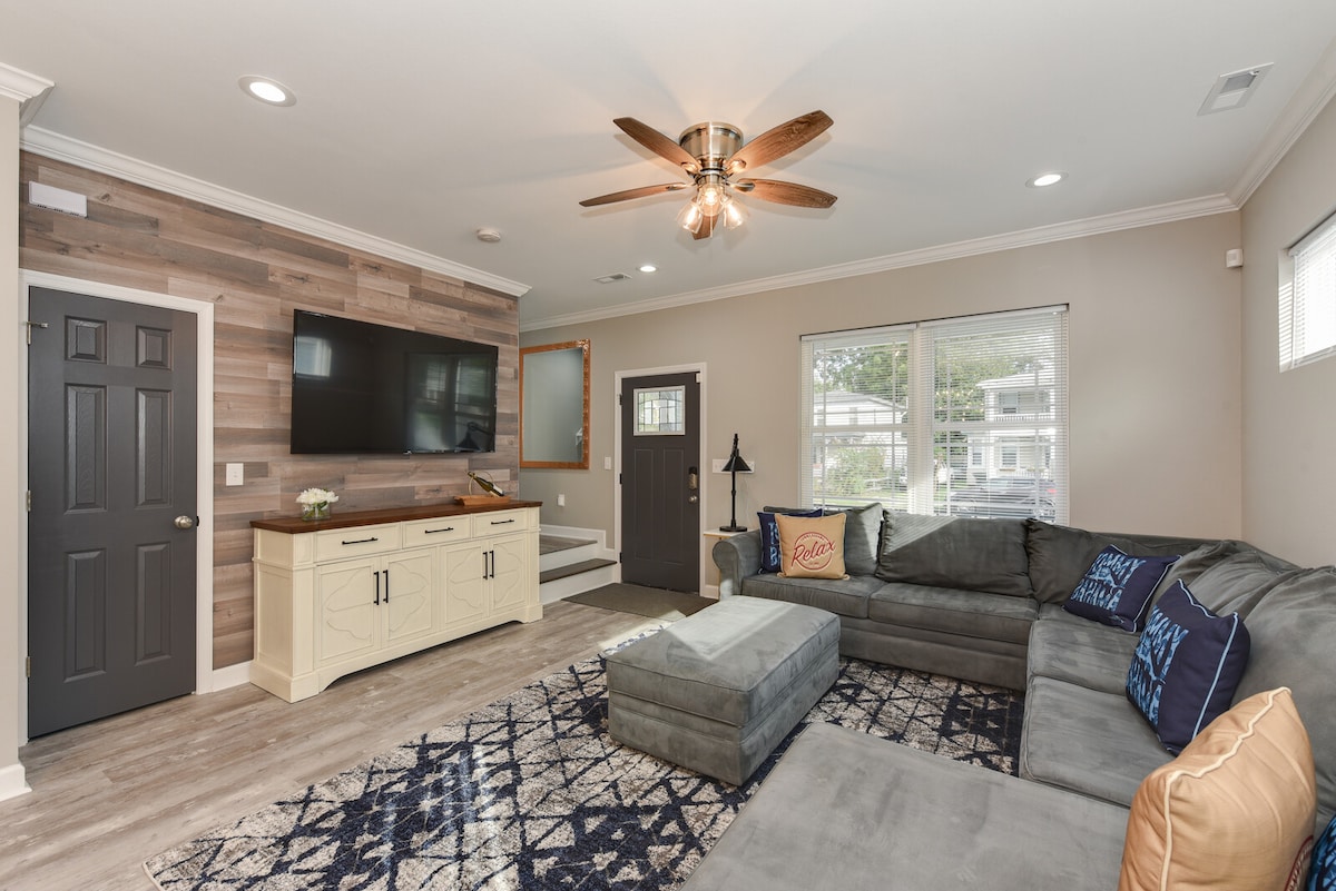 A cozy living room features a large sectional couch in shades of gray, complemented by a decorative blue and white area rug. A flat-screen TV is mounted on the wall, and a ceiling fan adds comfort. Natural light streams through the windows, enhancing the space.