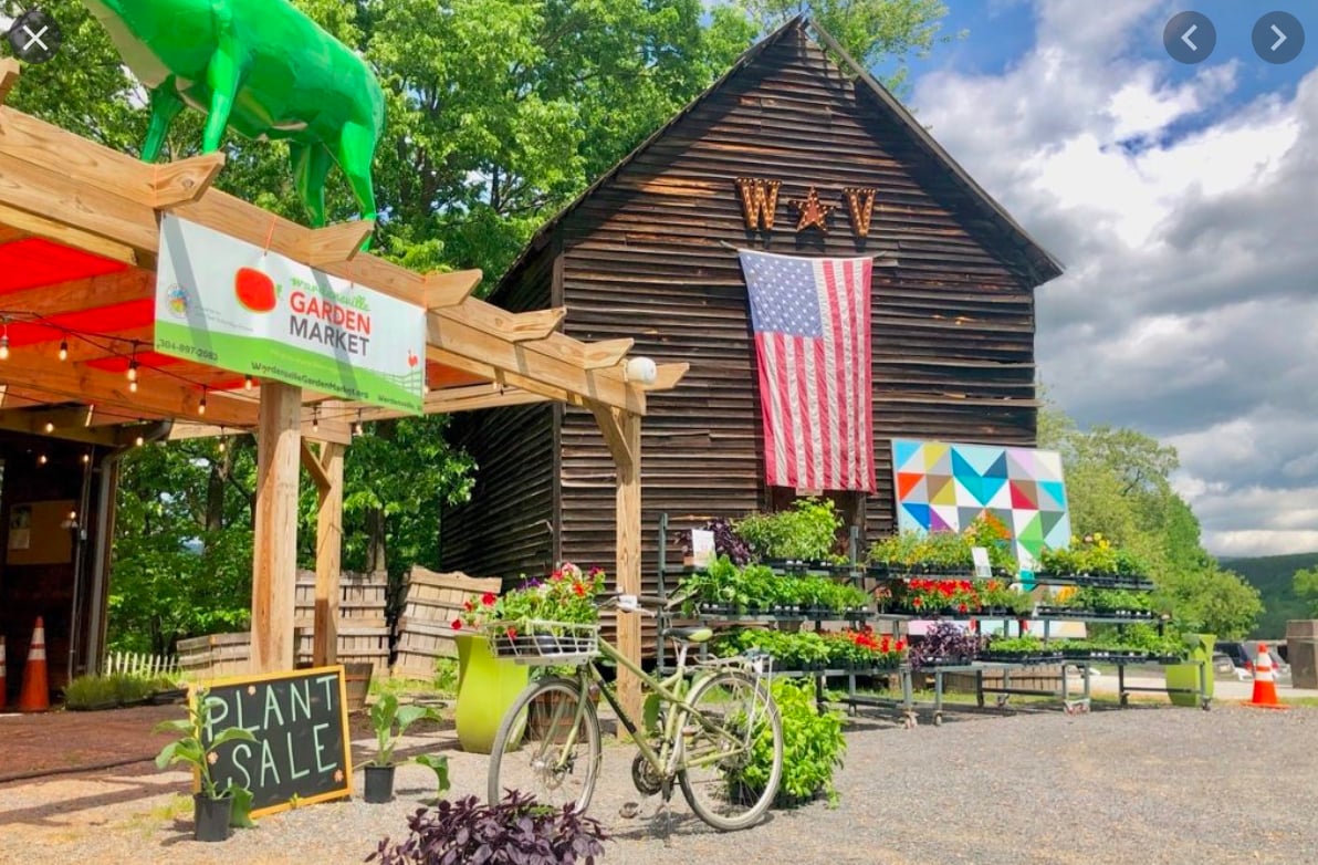 A rustic market building is adorned with a large American flag and features a vibrant sign announcing a plant sale. Potted plants and colorful flowers are displayed on stands outside, complemented by a vintage bicycle with a flower basket. Lush greenery surrounds the area.