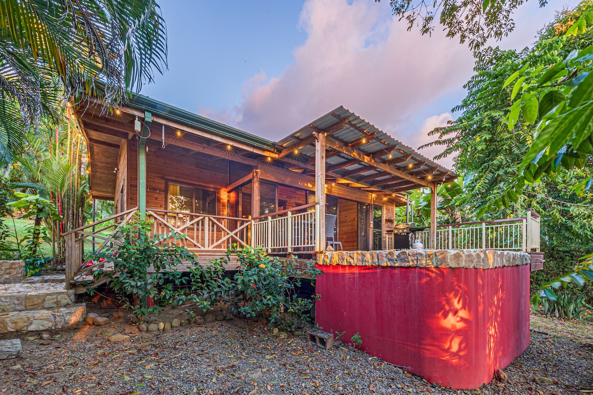 The cabin is surrounded by lush greenery and tropical plants, highlighting a rustic wooden design. A wraparound deck with railings offers outdoor seating, while a private pool is partially visible on the property. Warm light glows from the windows, inviting a sense of comfort.