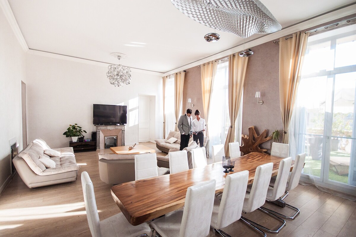 A spacious living and dining area features high ceilings and large windows allowing natural light to fill the room. A long wooden dining table accommodates several chairs, while soft furnishings and a television enhance the comfortable ambiance.