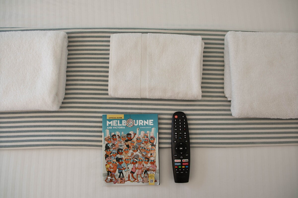 A neatly arranged bed display features three folded white towels positioned on a striped runner. A travel guide to Melbourne and Victoria is placed on the runner, alongside a remote control, indicating entertainment options within the room.
