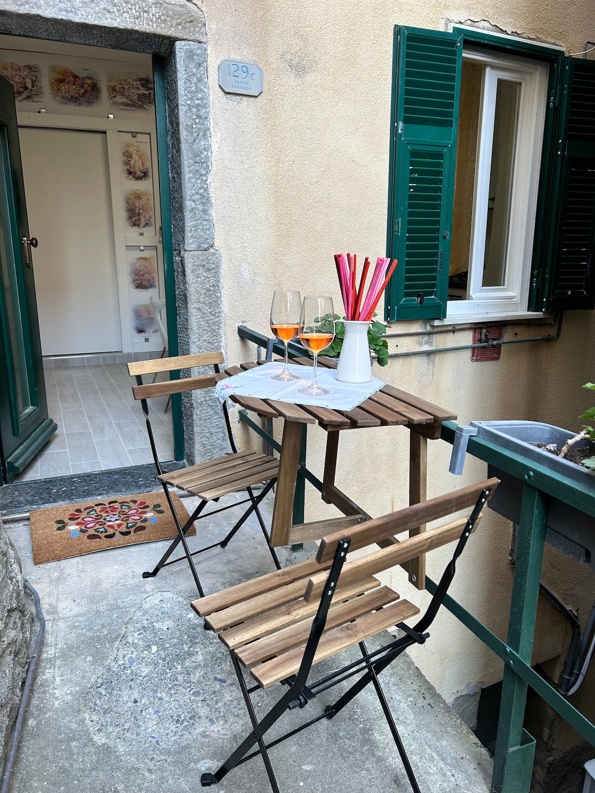 A small outdoor area is showcased with a wooden table and two folding chairs. A vase holds colorful straws, and two glasses sit on the table. The entrance to the loft is visible in the background, featuring bright green shutters and a welcoming mat.