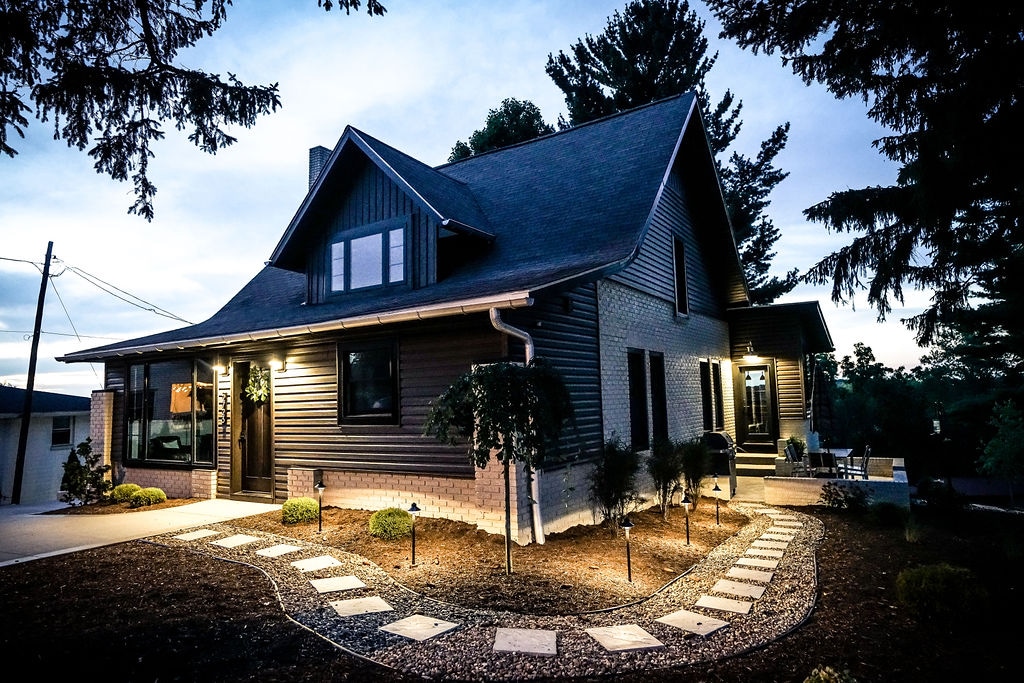 The exterior of the house is presented, showcasing a charming two-story design with a dark, wooden facade. Soft lighting highlights the intricacies of the architecture, while a path of stepping stones leads to the entrance. Lush greenery and a cozy seating area are visible.