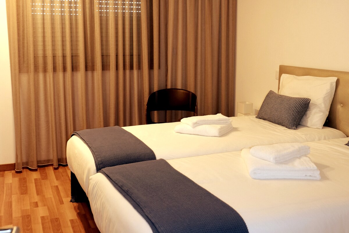 The sleeping area features two neatly arranged beds with crisp white linens and coordinating blue throws. A chair is positioned beside a window draped with light brown curtains, allowing for natural light to softly illuminate the space.
