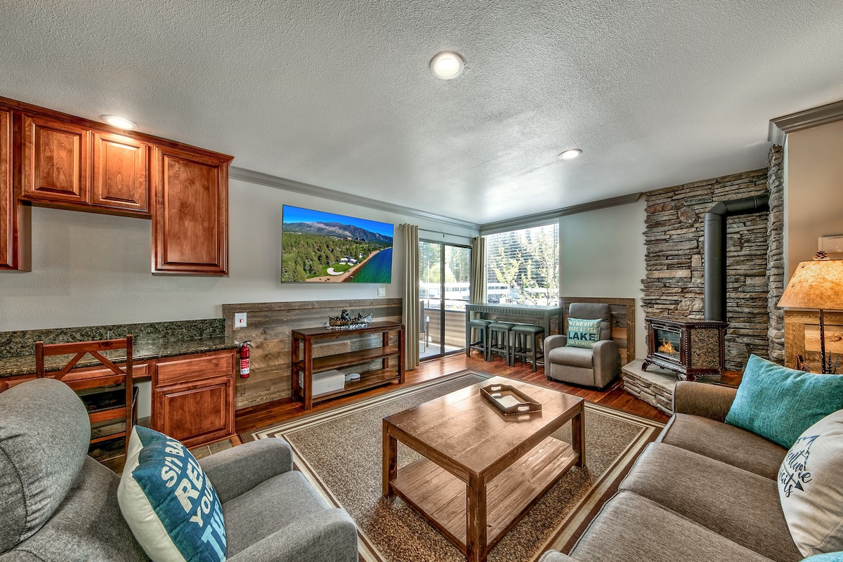 An open-concept living area is featured with a comfortable seating arrangement consisting of two sofas and two grey recliners. A wooden coffee table is centered on an area rug, and a stone fireplace is visible in the corner. Large sliding doors provide access to the outdoor balcony.