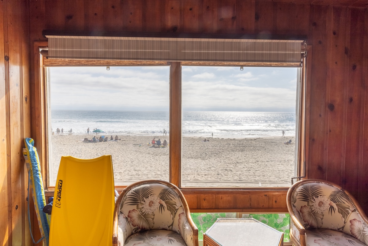 A spacious room features two large picture windows offering an unobstructed view of the beach and ocean. Soft sunlight reflects off the water, while beachgoers are seen in the distance. A yellow boogie board and rattan chairs are positioned nearby, enhancing the coastal ambiance.