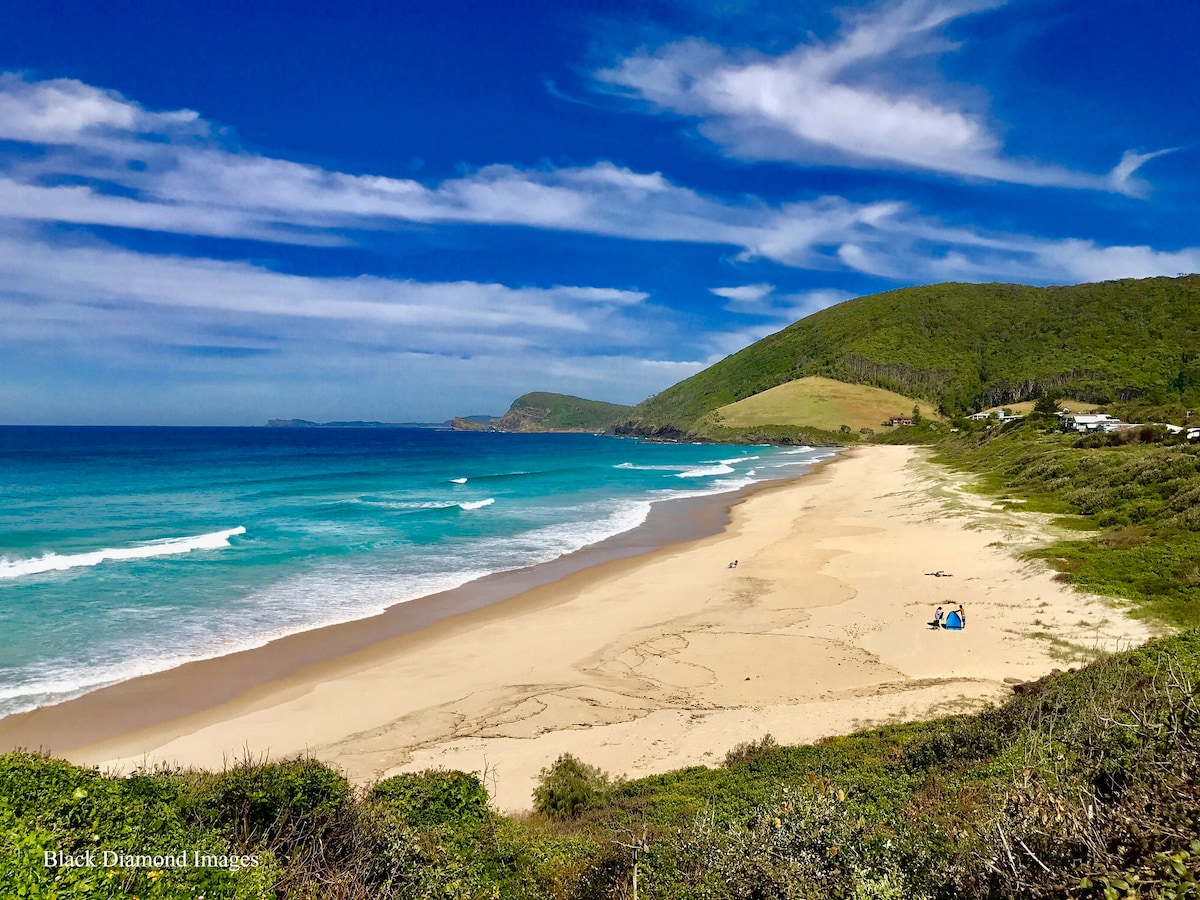 A panoramic view of Blueys Beach showcases soft golden sands meeting gentle turquoise waves. Lush greenery borders the beach, with rolling hills in the background. The sky is partially clouded, creating a serene atmosphere, inviting relaxation and outdoor activities.