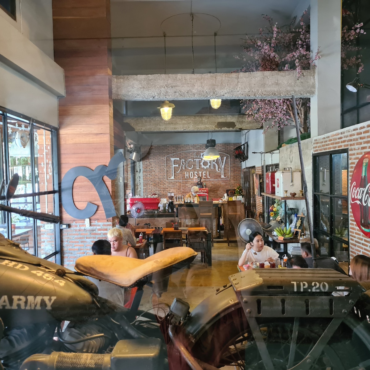 The spacious communal area is characterized by wooden accents and exposed brick walls, creating a welcoming environment. Tables and chairs are arranged for group seating, while a coffee counter is visible in the background. Natural light fills the space through large windows.