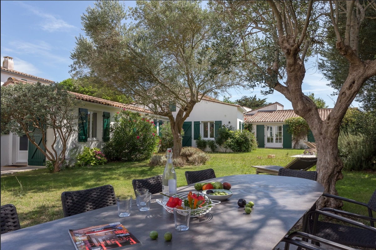 An outdoor dining area showcases a large table set with fresh fruits and beverages. Lush greenery surrounds the space, featuring a tree providing shade. The villa's exterior is visible, highlighting its windows and charming detailing against a clear blue sky.