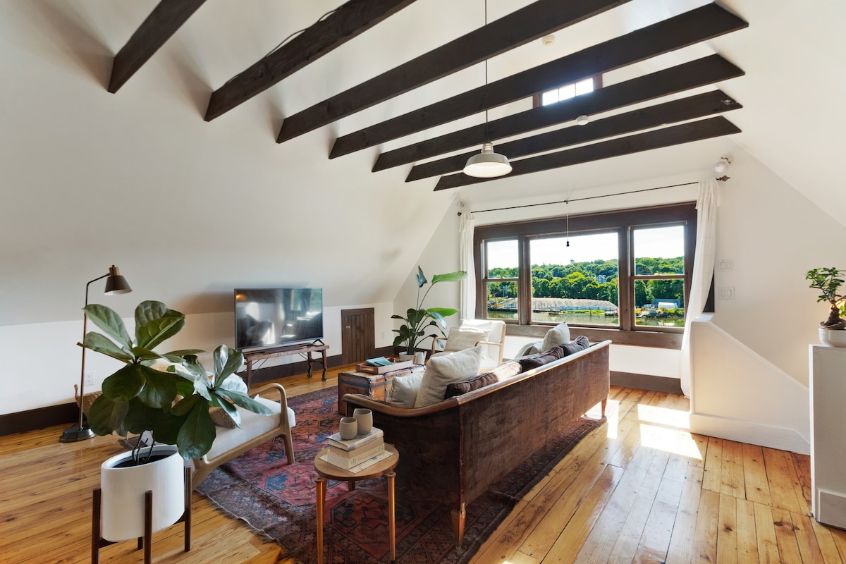 The spacious living area features high ceilings with exposed beams, large bay windows, and a comfortable seating arrangement. Natural light floods the room, highlighting the hardwood floors and greenery from a potted plant. A television is positioned beside a wooden cabinet.