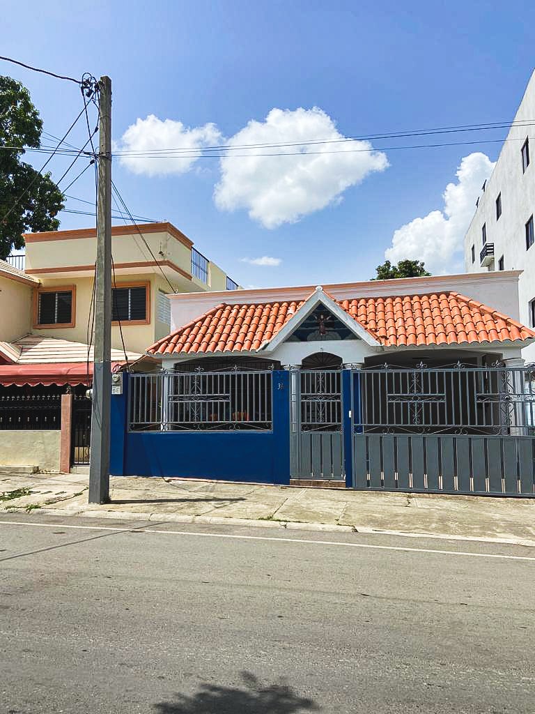 The exterior of a charming house is visible, featuring a red-tiled roof and blue accents. Metal bars decorate the windows and doors, providing security. A small front yard is bordered by a low fence. Clear blue skies with scattered clouds create a pleasant backdrop.