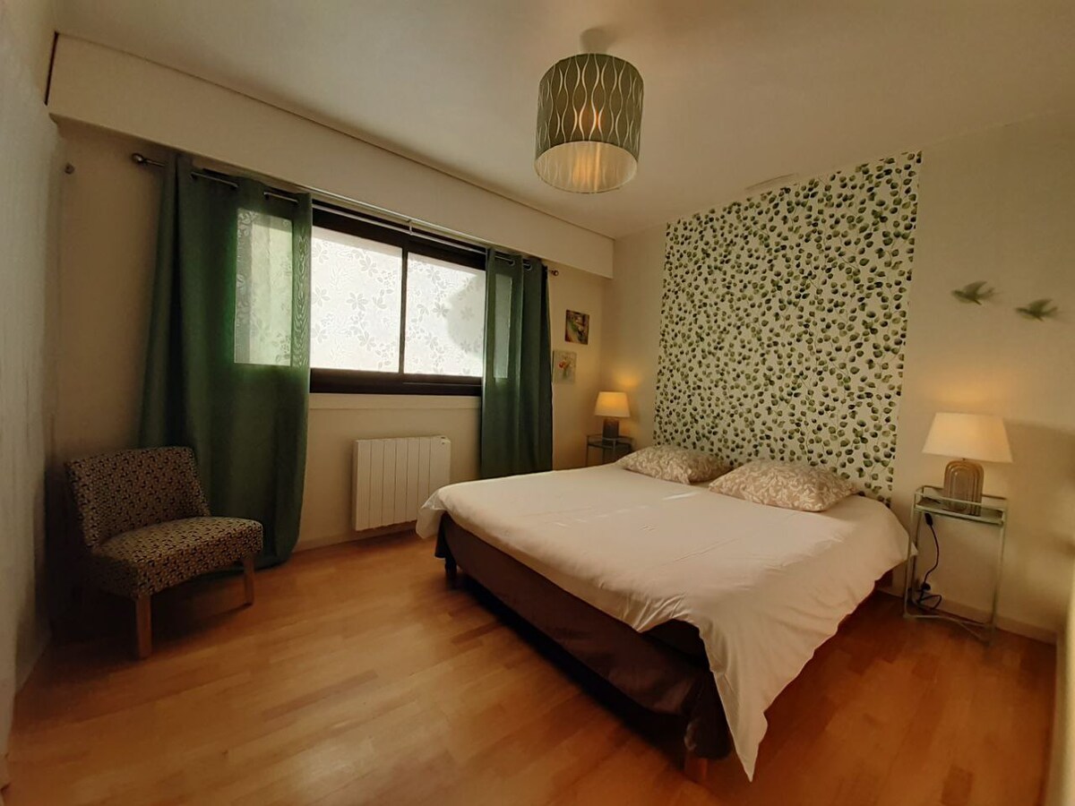 A spacious bedroom is shown, featuring a large bed with light linens and two decorative pillows. A patterned feature wall complements the room. Natural light filters through a window dressed with green curtains. A small armchair and bedside lamps add to the welcoming ambiance.