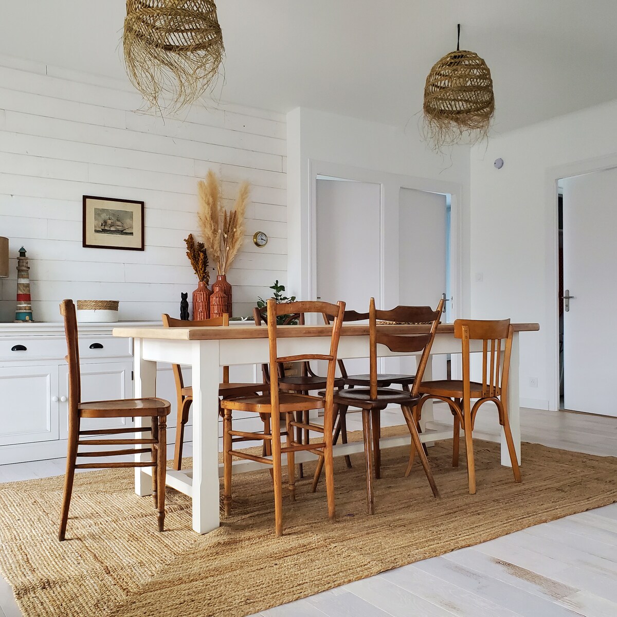 A dining area is presented with a large white table surrounded by wooden chairs. Two woven pendant lights hang from the ceiling, and a textured rug covers the floor. Decorative vases and dried plants add subtle detail to the space.