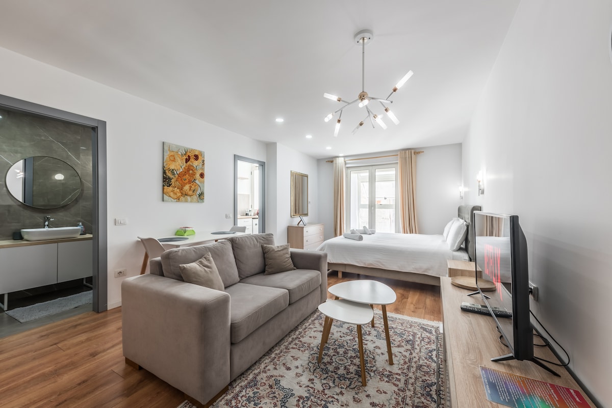 A spacious studio apartment features a comfortable double bed with soft linens and a modern TV unit equipped with a 43" Smart TV. A cozy sofa is positioned beside a small coffee table. Natural light filters through large windows, illuminating the stylish interior.