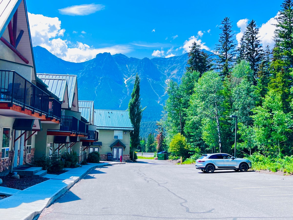 A scenic view captures the two-story townhouses with a backdrop of towering mountains and a bright blue sky. Lush greenery lines the parking area, while a silver car is parked nearby. The layout of the complex allows for spacious outdoor areas.