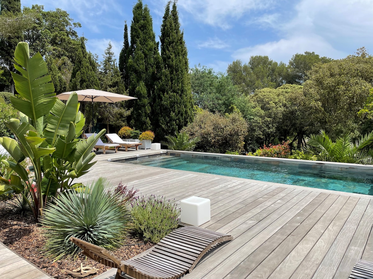 A serene outdoor space features a salt-water pool surrounded by wooden decking. Lush greenery, including tall trees and tropical plants, enhances the private garden atmosphere. Comfortable lounge chairs with an umbrella offer shade, contributing to a tranquil and inviting environment.