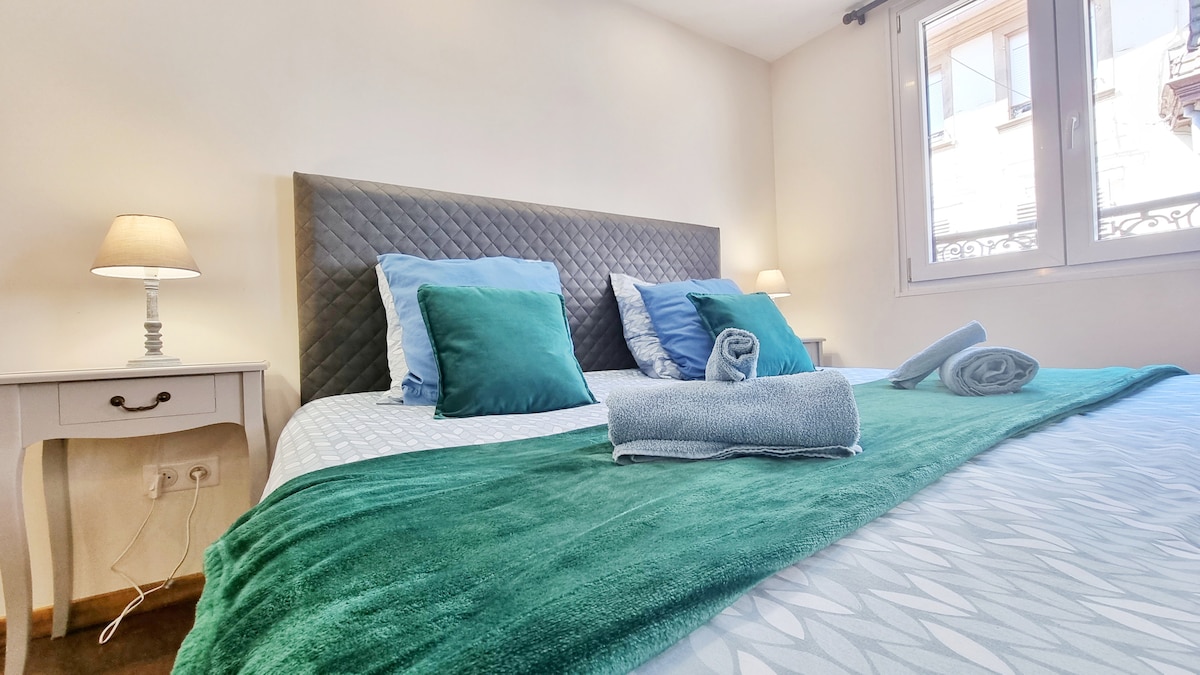A spacious sleeping area features a plush bed adorned with decorative pillows and a soft blanket in calming green tones. Two side lamps provide gentle illumination beside a nightstand. Natural light enters through a window, enhancing the inviting ambiance of the room.