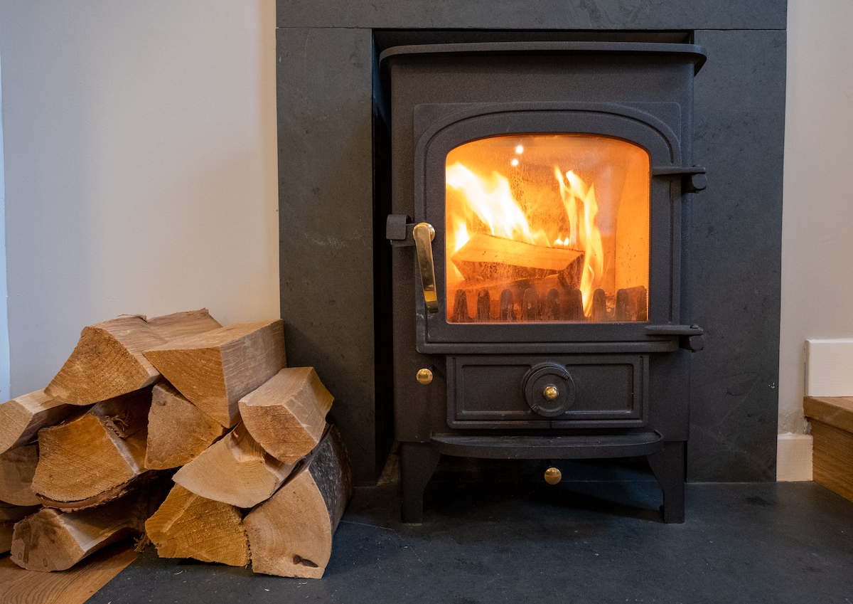 A wood-burning stove emits a warm glow, surrounded by neatly stacked logs. The stove is set within a stone hearth, offering a cozy focal point for the room.