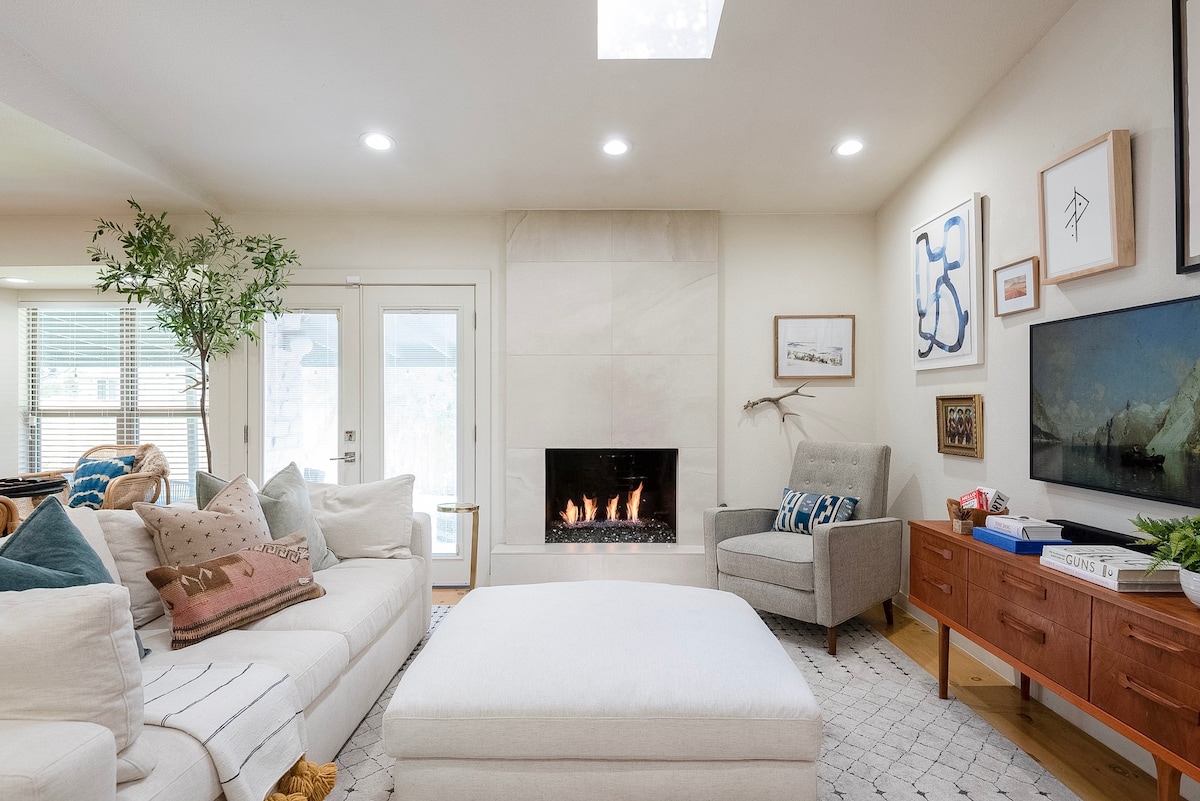 A bright living room is featured, highlighting a cozy deep sofa adorned with textured pillows and a large ottoman. A modern fireplace stands against a sleek stone backdrop, while natural light filters through windows, enhancing the warm ambiance of the space.