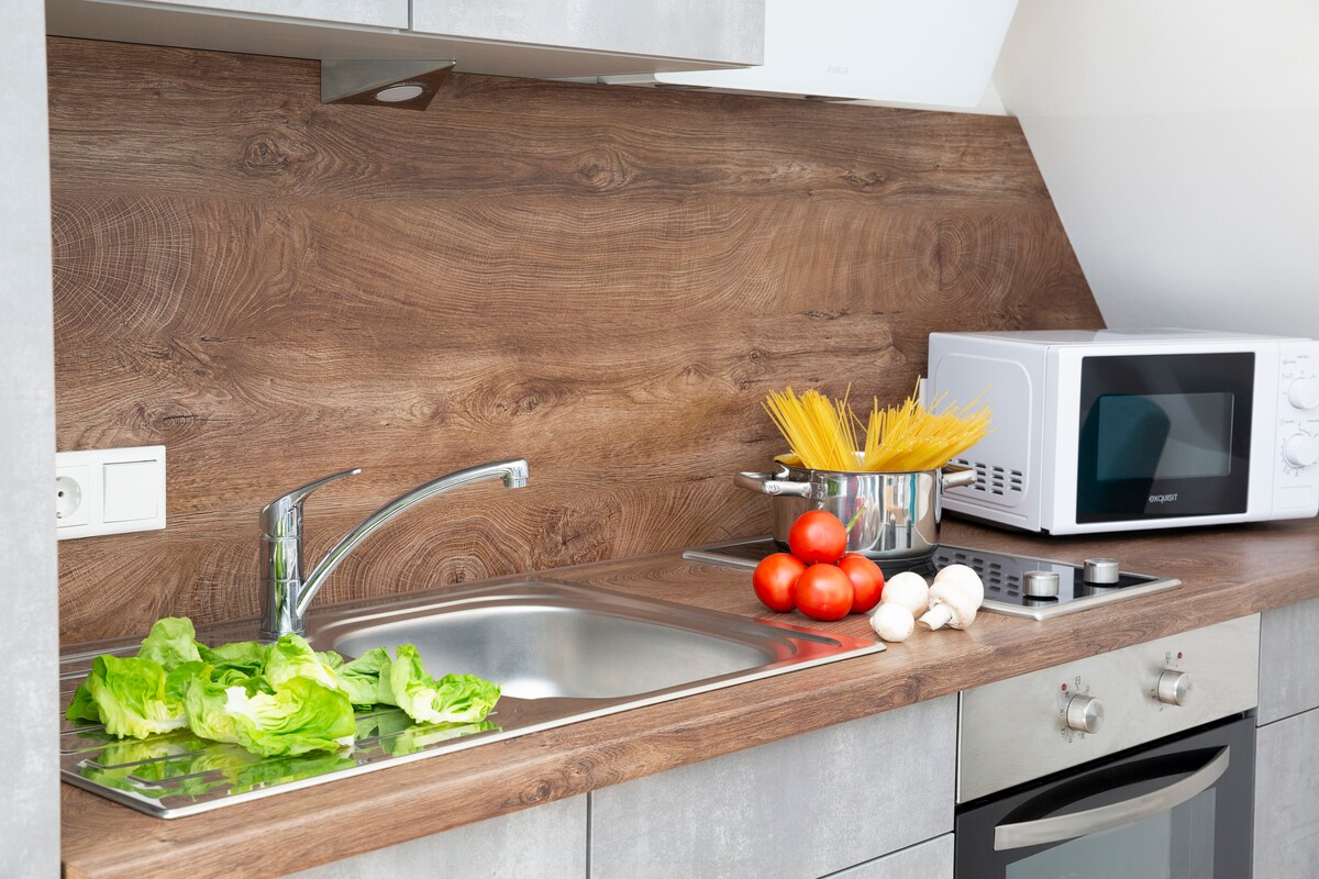 A modern kitchen space is presented, featuring a stainless steel sink with fresh lettuce. A cutting board is nearby, with tomatoes and mushrooms placed beside a pot of spaghetti. A microwave and stove are visible, accentuated by warm wooden paneling.