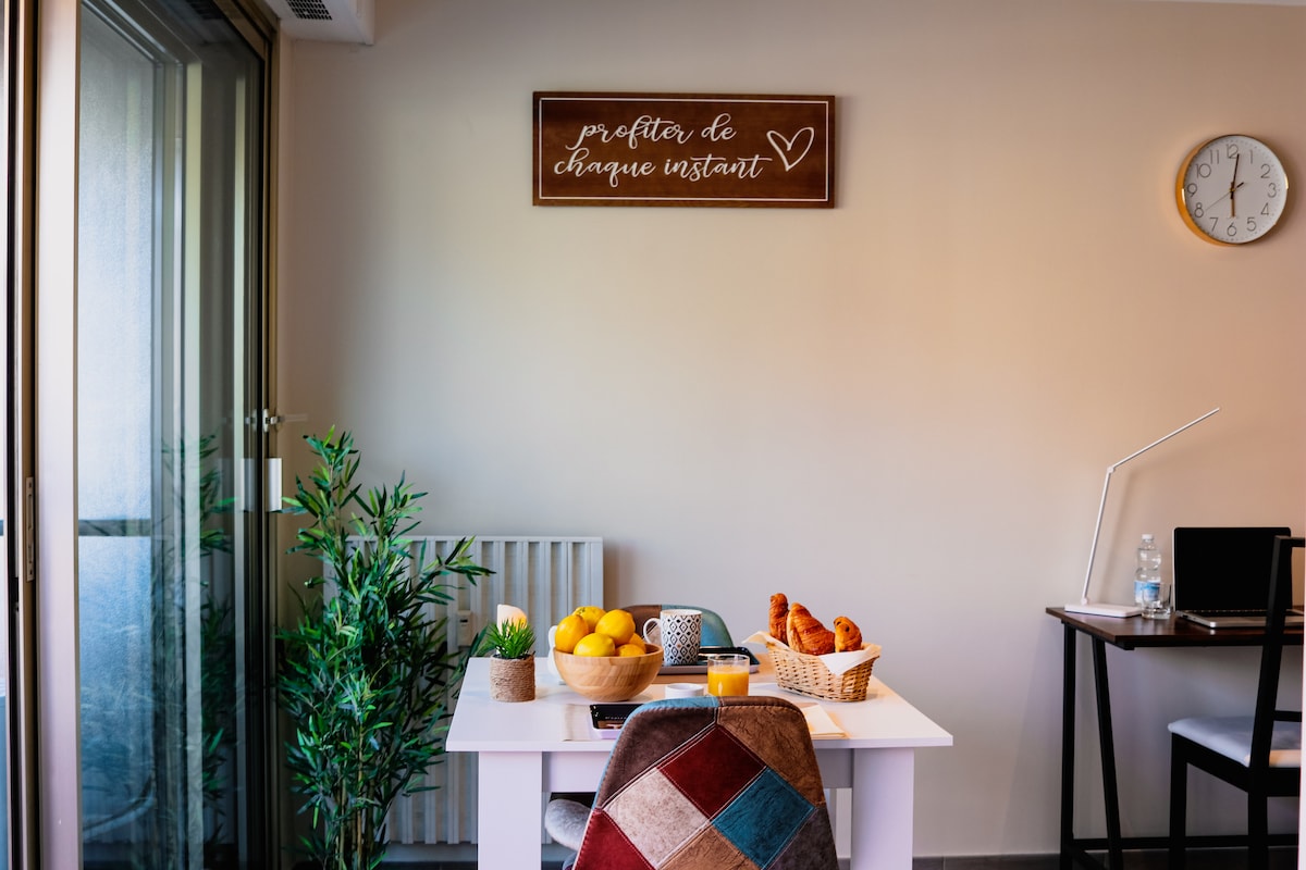 A dining area is seen, featuring a white table adorned with a basket of croissants and a bowl of fresh fruit. A small plant adds a touch of greenery, while a wall clock and a decorative wooden sign complete the space. Natural light flows through the nearby sliding glass doors.