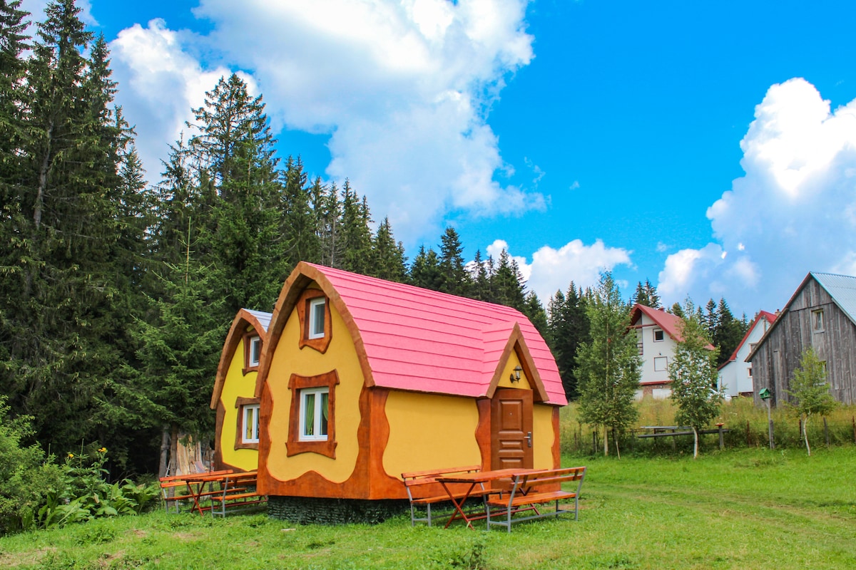 A whimsical bungalow is set amidst a lush green landscape, featuring a vibrant yellow exterior with reddish-brown accents and a distinctive red roof. Surrounded by tall trees, two wooden picnic tables are positioned in front, offering a pleasant outdoor seating area.
