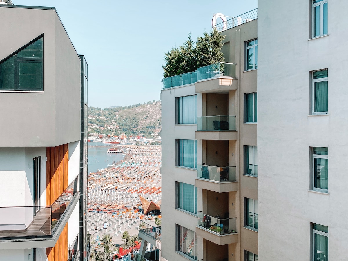 The image displays a view between two buildings towards a vibrant beach area. Multiple layers of colorful beach umbrellas line the shore, with a mountainous backdrop in the distance. Balcony spaces are visible on the right building, enhancing the scenic ambiance.