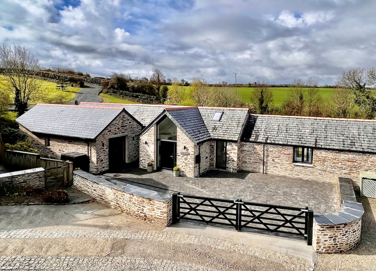 The exterior of the barn conversion is showcased, featuring natural stone walls and a slate roof. A spacious driveway is visible, with a gated entrance leading to the house. Lush green fields stretch in the background under a partly cloudy sky.