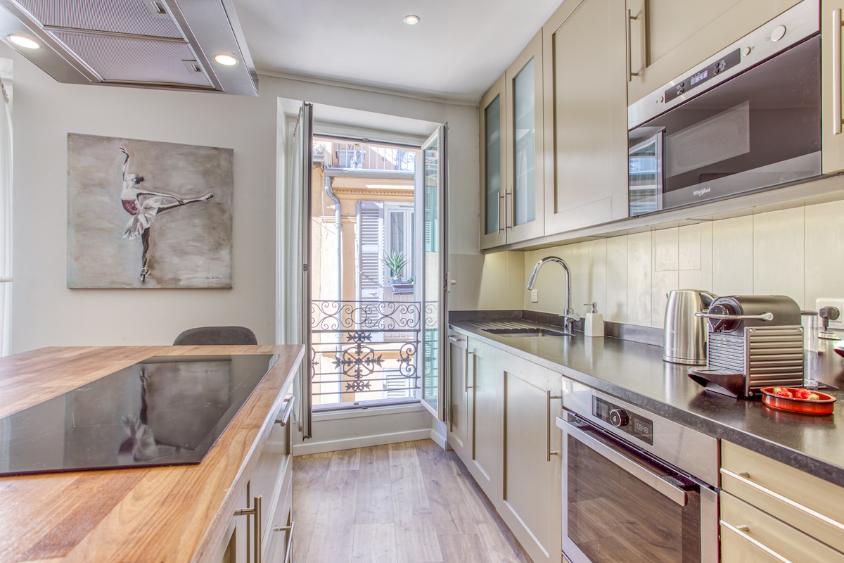 A modern kitchen featuring sleek cabinetry with a beautiful countertop and a central island. Large windows open to a charming balcony, allowing natural light to flow. State-of-the-art appliances, including a kettle and microwave, are neatly arranged along the counters.