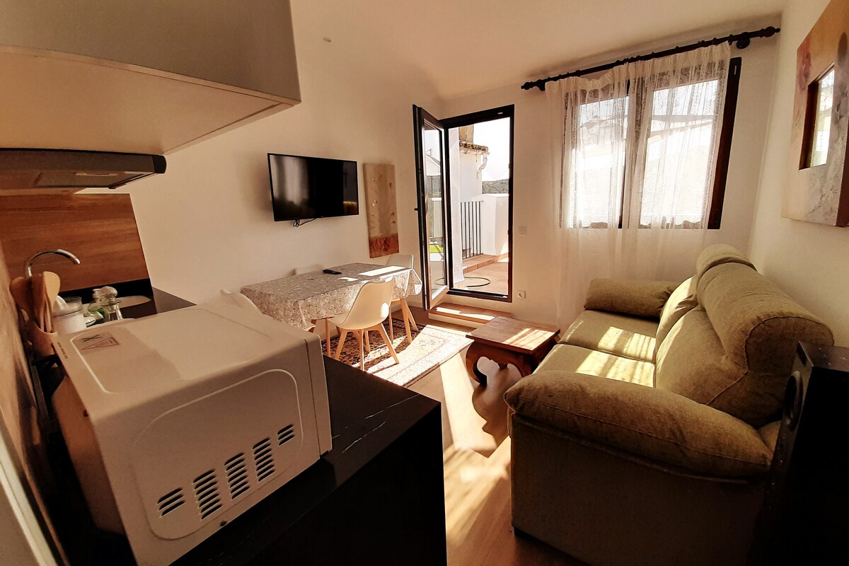 A well-lit living area showcases a cozy green sofa and a wooden coffee table. A dining space with white chairs is visible, accompanied by a television mounted on the wall. Large glass doors open to a private outdoor area, inviting natural light inside.