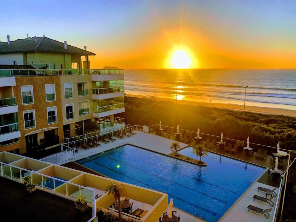 A breathtaking sunset casts a warm glow over the ocean, visible beyond a spacious outdoor pool. The scene is framed by a well-maintained building featuring multiple balconies, and sun loungers are arranged by the water's edge, inviting relaxation.