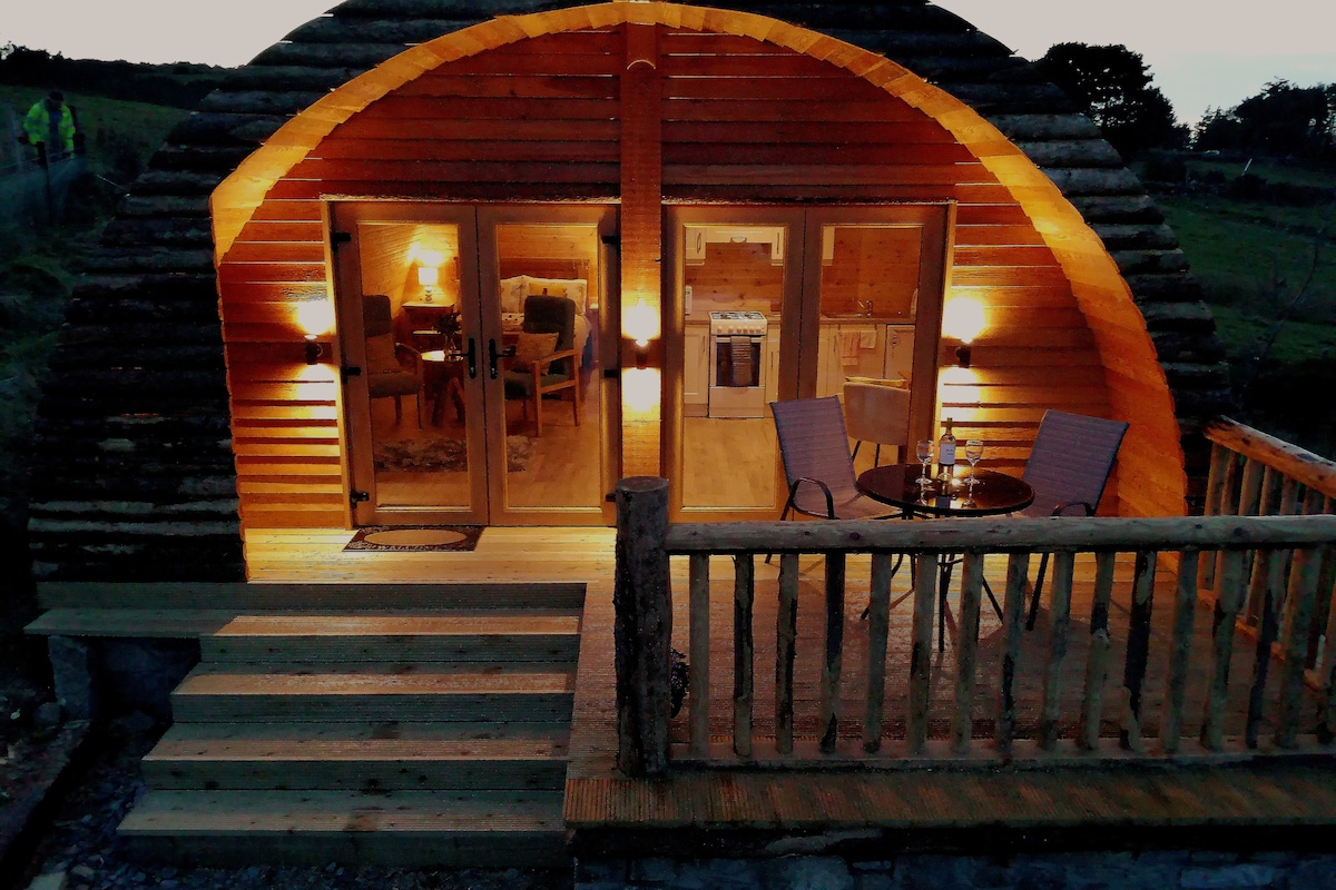 A log cabin exterior is illuminated by warm lights during twilight. The entrance features a wooden deck with a table and chairs, inviting relaxation. Glass doors provide a glimpse into the cozy interior, showcasing a living area with soft lighting and modern appliances.