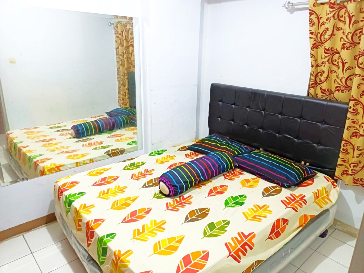 The sleeping area features a mirrored wall that reflects a spacious bed dressed in a colorful leaf-patterned blanket. Two striped pillows are placed at the headboard, creating a vibrant contrast against the neutral decor. Light-colored curtains frame the window, adding to the bright ambiance.