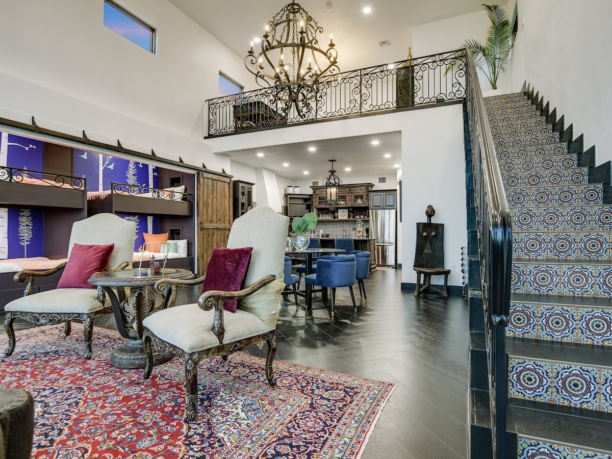 A spacious living area showcases intricate tile flooring and high vaulted ceilings supported by wrought iron railings. Elegant furnishings, including two armchairs with plush pillows, are arranged around a decorative rug. A large chandelier provides soft illumination, enhancing the warm atmosphere.